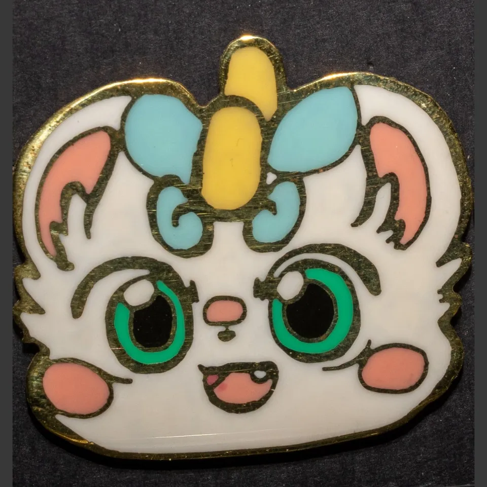 Enamel pin of a white cat with a yellow horn, blue bow and big green eyes.