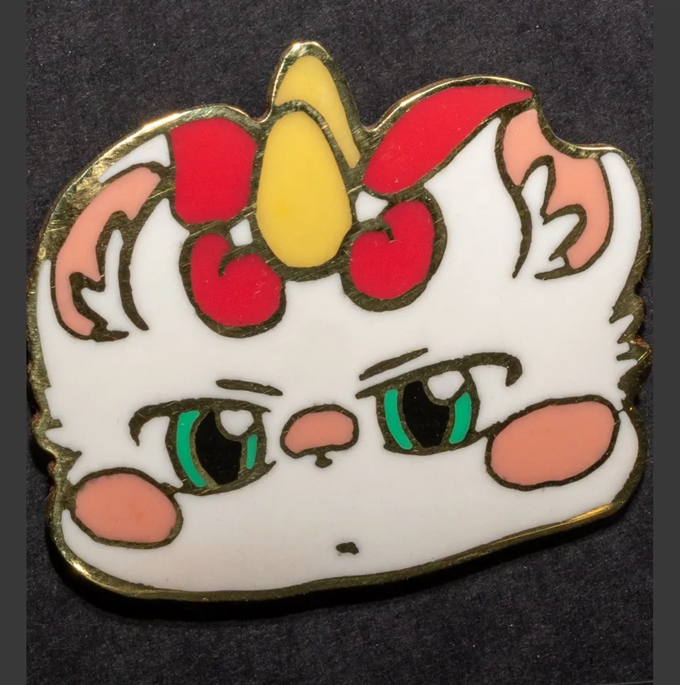 Enamel pin of a white cat with a yellow horn, red bow and a grumpy face.